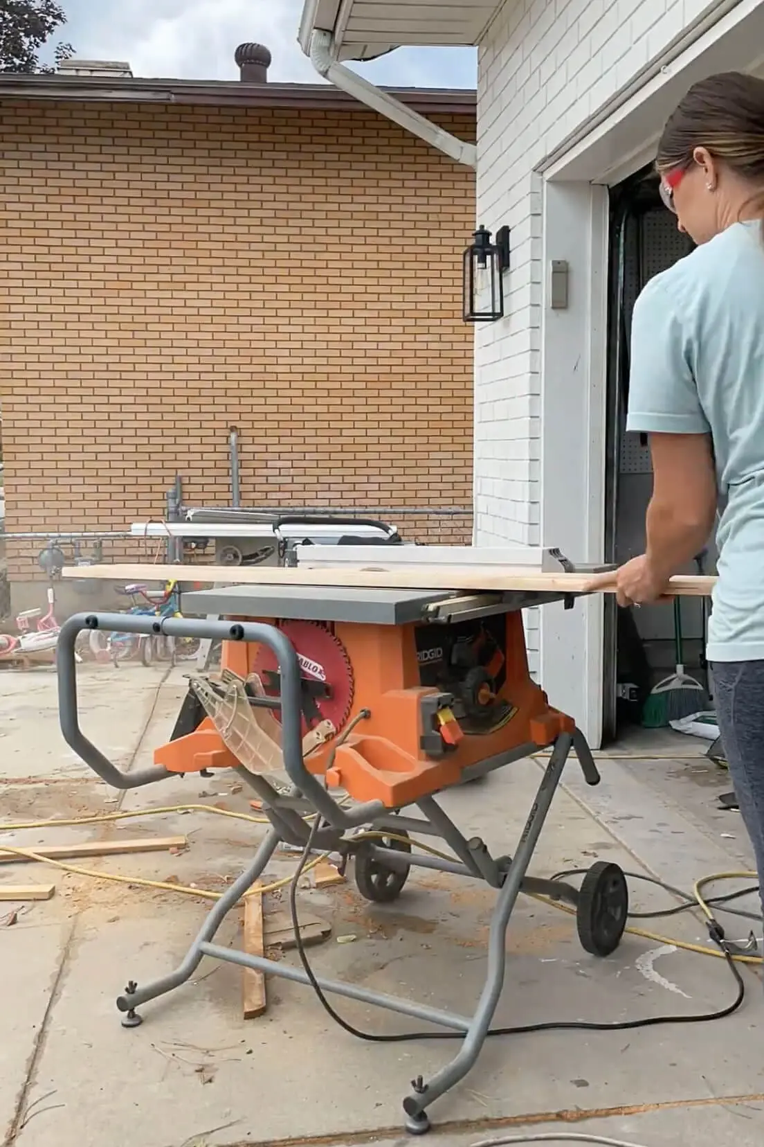 Woman using a Ridgid portable table saw to cut lumber for a DIY project, surrounded by sawdust and woodworking tools on a driveway workspace.